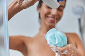 Cheerful young woman pouring shower gel on bath loofah