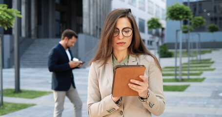 Fototapeta premium Caucasian attractive young stylish woman tapping or scrolling on tablet device in town at street. Beautiful female texting message and using gadget computer. Businesswoman in glasses working outdoor.