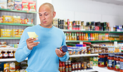 Positive male customer with shopping list choosing food products in supermarket