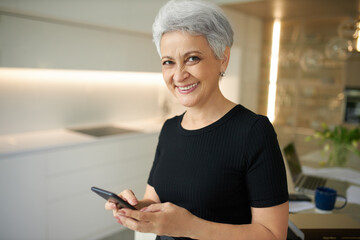 Electronic gadgets and communication concept. Cheerful positive middle aged female with short hairdo messaging with friends online via group chat on cell phone, inviting them for dinner