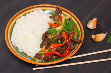 Roast beef with sweet pepper and white rice. A plate with colored stripes, garlic cloves, dark background and Chinese wooden chopsticks.