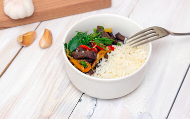 Roast beef with sweet pepper and white rice. White plate, garlic in the background. Side view. Delicious dish.