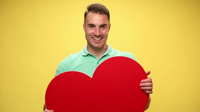 handsome casual guy raising his big red heart and showing it at the camera on yellow background