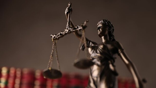 bronze law statue represented by blindfolded goddess spinning and holding a balance signifying impartiality and two broad sources of law in front of books on brown background