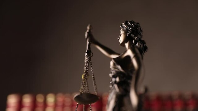 down view of justice statue represented by blindfolded lady holding a balance and a sword signifying power and impartiality, rotating in front of red books on brown background