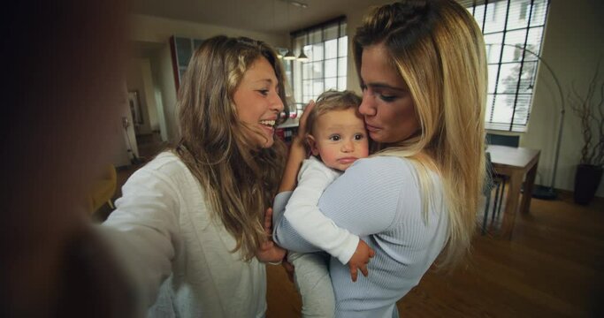 Authentic shot of young happy homosexual female gay family with son toddler baby boy is making a selfie or video call to friends or relatives in a living room at home.