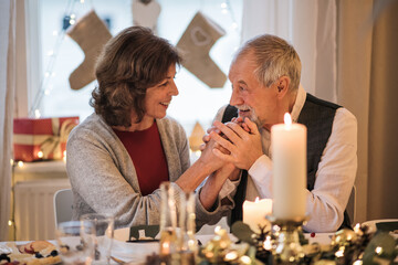 Happy senior couple indoors at home sitting at the table at Christmas, talking.
