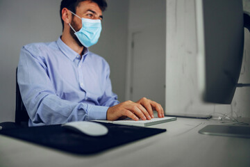 Young attractive businessman with protective mask on sitting in office and typing report.