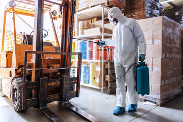Fototapeta na wymiar Worker in sterile uniform with rubber gloves holding sprayer with disinfectant and spraying around warehouse. Corona outbreak concept.