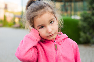 A little girl in a pink hoodie with a sad and tearful face is holding her ear. Ear pain, otitis...