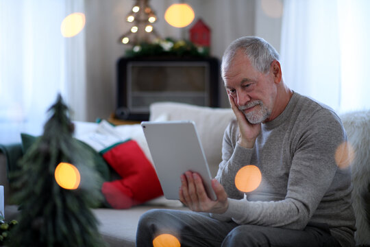 Sad And Lonely Senior Man With Tablet Sitting At The Table Indoors At Christmas, Solitude Concept.