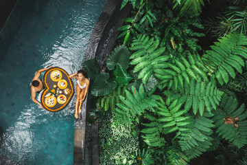 Travel happy couple in love eating floating breakfast in jungle swimming pool. Awakening in morning. Black rattan tray in heart shape, Valentines day or honeymoon surprise, view from above. - 382985550