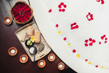 Romantic spa set with lime, scrub, salt, towels and brush in wooden tray. Flower petals in bath tub with foam and candles around. Organic and natural beauty treatment accessory. 