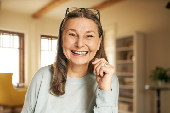 Close up image of happy beautiful energetic senior woman with wrinkles and gray long hair enjoying nice time indoors posing in cozy living room interior, looking and smiling broadly at camera