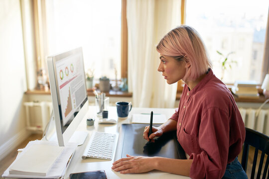 Talented stylish young woman illustrator with pink bob hairdo holding cup drinking coffee while working on visual content for website, using graphic tablet, hand drawing images or animation