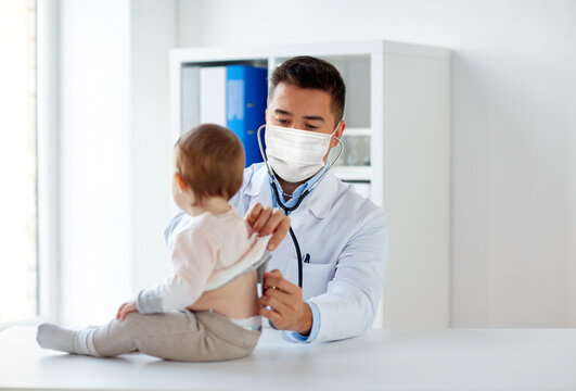 Medicine, Healthcare, Pediatry And People Concept - Doctor With Stethoscope Wearing Face Protective Medical Mask For Protection From Virus Disease Listening To Baby On Medical Exam At Clinic