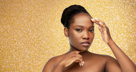 Obraz na płótnie Canvas beauty, luxury and people concept - portrait of young african american woman with bare shoulders touching her face over golden glitter background