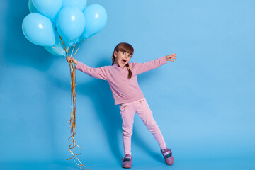 Fototapeta na wymiar Girl with bunch of helium balloons spreading hands aside and yelling something happily, wearing casual rosy jumper, trousers and sneakers against blue background, cute child at party.