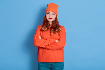 Good looking young European woman being calm, keeps arms folded, wears orange knitted sweater and cap, stands against blue background, red haired female looks at camera.