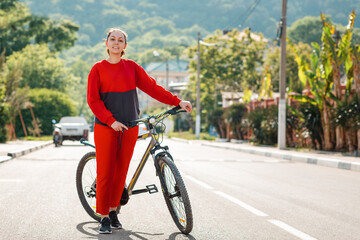 Naklejka premium Sports and activity. A pretty woman in red sportswear is pushing a Bicycle along an empty road. Copy space