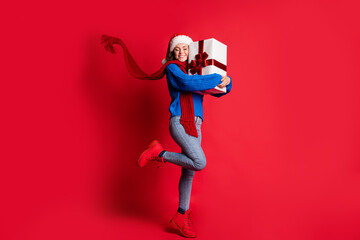 Photo portrait full body cheerful woman hugging present box smiling closed eyes standing on one leg scarf flowing isolated on bright red colored background