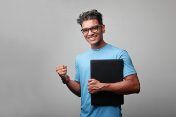 Smiling young man of Indian origin holding a laptop in his hand with a winning gesture