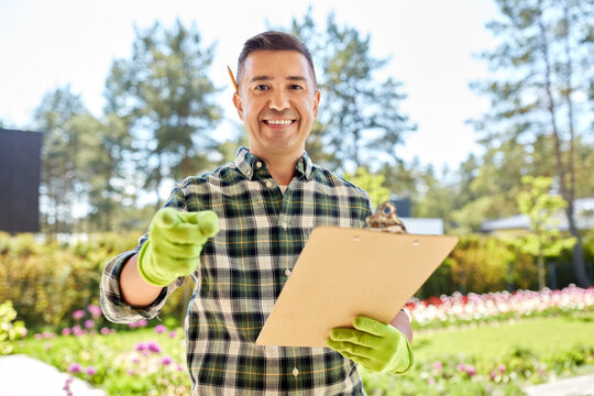 gardening and people concept - happy smiling middle-aged man in apron with clipboard pointing to camera at summer garden