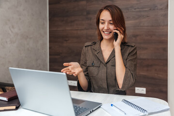 Happy young woman talking on mobile phone and looking at laptop