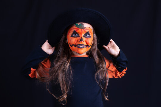 smiling girl with pumpkin makeup on her face holding a black hat on a black background, funny Halloween celebration.