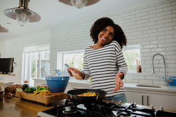 Smiling ethnic woman chatting on smartphone while holding spatula making lunch in pan standing in modern kitchen.