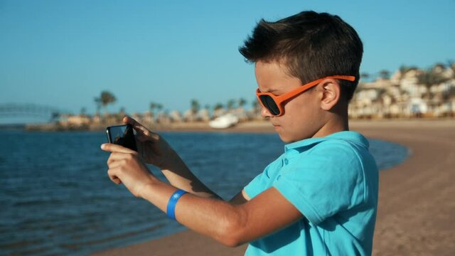 Attractive teenage boy taking pictures by mobile phone at sunny coastline.