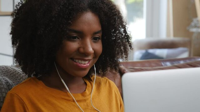 Beautiful girl with afro wearing earphones listening to online lecture on laptop sitting on sofa in lounge.