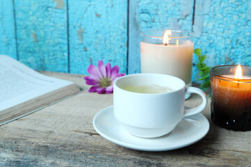 a white cup of ice jasmine tea is on the wooden table with a book and two glass scented aroma candles on the background of vintage blue wooden wall of the house during tea time in the afternoon 