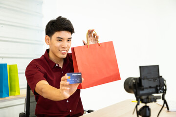 Young Asian male online merchant blogger using camera to live video, show shopping bag and credit card. Social media, Influencer, online shopping, and payment concept.