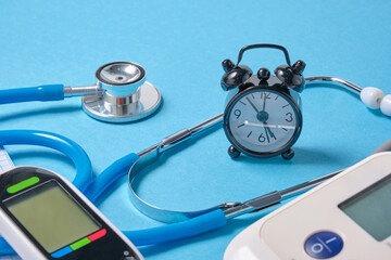 Digital blood pressure monitor, glucometer, stethoscope, notepad and alarm clock on blue background