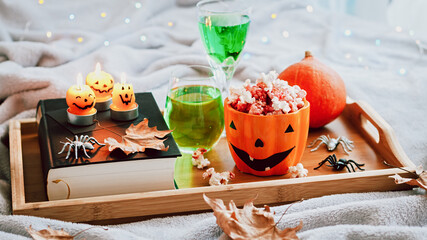Book, halloween decor, candles, popcorn and drinks on tray on bed, halloween reading concept with bokeh background. Still life for Halloween at home