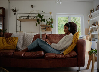 Obraz premium Side view of young woman relaxing at home watching webinar and typing with headphones sitting on couch