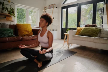 Abwaschbare Fototapete Young african woman meditating sitting in lotus position at home on yoga mat © StratfordProductions