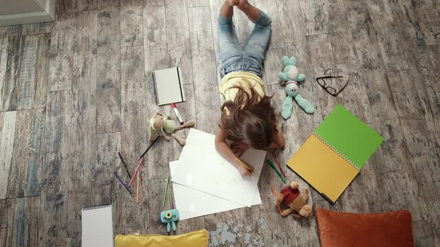 Concept of creativity. Top view of little girl lying on the wooden floor and drawing with colorful pencils on a white sheet of paper, playing alone at home