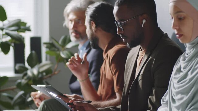 Tilt up shot of young Afro-American man in wireless earphones and smart casual outfit sitting with multiethnic candidates, filling application before job interview, then looking at camera and smiling