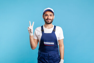 Optimistic handyman in overalls and hygiene gloves showing victory sign, profession of service industry, courier delivery, housekeeping maintenance. Worker in uniform gesturing like. studio shot