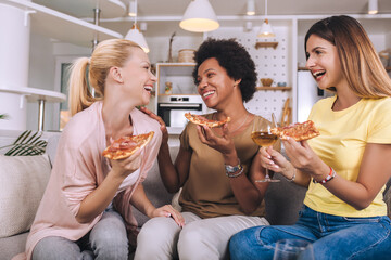 Happy young female friends with wine and pizza on sofa at home.