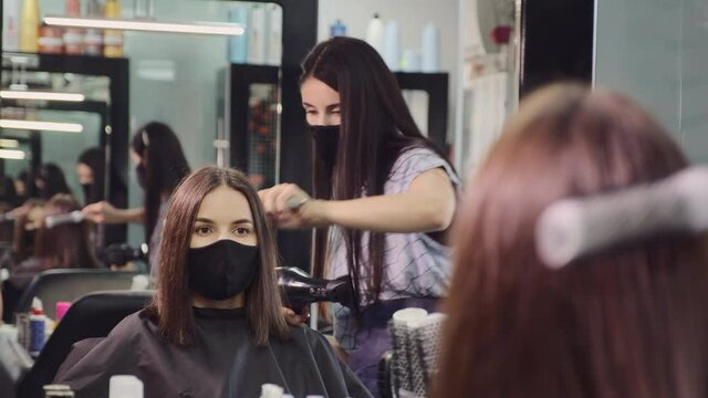 Woman stylist in black protective mask works with female client short hair using dryer by large mirror with replicated reflection in salon