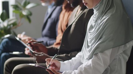Tilt down selective focus shot of young muslim woman in hijab sitting with diverse candidates and filling application form before having job interview