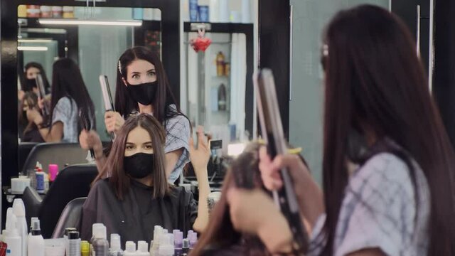 Joyful young hairstylist and woman client in protective masks dance having fun at large mirror with replicated reflection in beauty salon