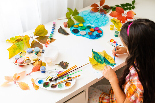 little girl paints leaves with brush at home, autumn art.