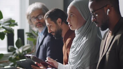 Tilt up shot of young muslim woman in hijab sitting with diverse male candidates in office, filling application form and then posing for camera while waiting for job interview