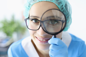 Young woman doctor in glasses and uniform holds magnifying glass near her eyes portrait....