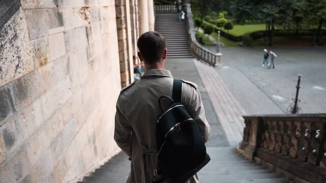 Slow motion shot of a young adult man wearing a trench coat and backpack walking down the stairs