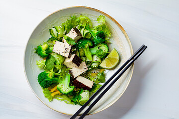 Green Asian salad with broccoli and smoked tofu in white bowl, white background.